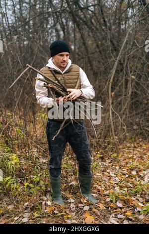 Vertikales Porträt eines gefrorenen Touristen-Mannes, der warme Kleidung trägt und trockenes Totholz für Feuer im Wald an einem bewölkten, kalten Tag sammelt. Stockfoto