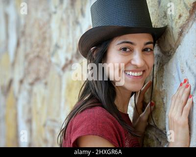 Die junge, schöne Brünette mit langen Haaren und roten Fingernägeln trägt einen schwarzen Zylinderhut und blickt auf den Betrachter vor einem Steinwandhintergrund. Stockfoto