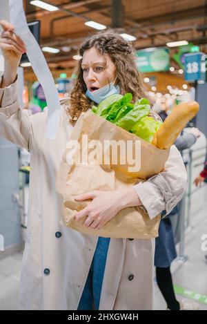 Die junge Frau ist unangenehm überrascht, als sie in einem Supermarkt in einen Papierscheck schaut und eine Einkaufstasche hält. Steigende Lebensmittelpreise. Stockfoto