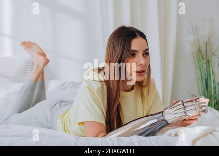Bloggerin mit einem bionischen prothetischen Arm leitet eine Online-Übertragung oder zeichnet ein Problem auf oder kommuniziert per Videoanruf mit Freunden, während sie an einem sonnigen Tag in bequemer Heimkleidung auf einem Bett liegt. Stockfoto