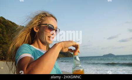 Frau trinkt am Strand Whiskey mit Orangensaft aus einer Flasche, entspannen Sie sich bei einem warmen goldenen Sonnenuntergang. Frau, die in blauem Badeanzug und Sonnenbrille läuft. Das Konzept ist gut Stockfoto