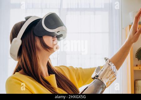 Positive junge Frau in legerer Kleidung mit modernem bionischem Arm und Virtual-Reality-Headset tanzt neben dem Sofa in der stilvollen Küche Stockfoto