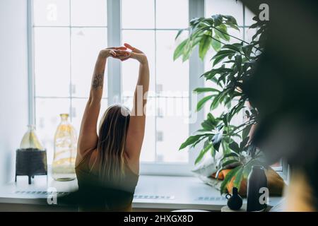 Frau mit blonden langen Haaren, die ein leuchtend gelbes Sporttop trägt, sitzt auf dem Bett mit weichen Kissen und blickt aus dem Fenster Stockfoto