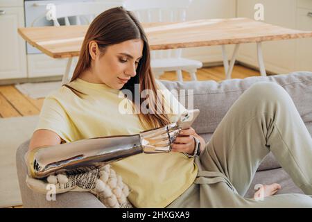 Eine glückliche Frau mit einem bionischen prothetischen Arm sitzt mit einem Telefon auf dem Sofa und kommuniziert online mit Freunden oder surft im Internet oder kauft online in einem gelben T-Shirt vor dem Hintergrund einer hellen Küche ein Stockfoto