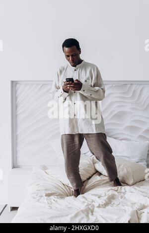 Ein lustiger, aufgeregter junger afroamerikanischer Mann in legerer Kleidung springt in einem modernen Zimmer mit Mobiltelefon auf einem großen Bett hoch Stockfoto