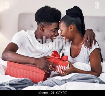 Austausch ihrer ersten Jubiläumsgeschenke. Aufnahme eines jungen Paares, das Geschenke austauschte, während es auf ihrem Bett saß. Stockfoto