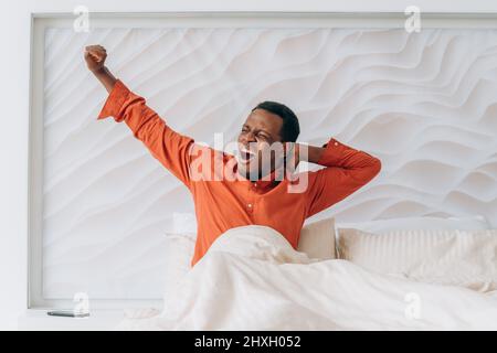 Der junge afroamerikanische Mann im orangefarbenen Pyjama gähnt und streckt sich in einem bequemen Bett in der Nähe des frühen sonnigen Morgens auf Stockfoto