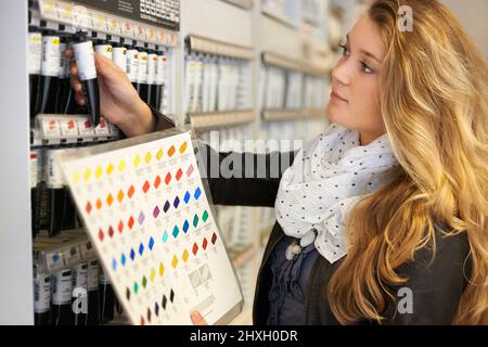 Qualität statt Preis. Aufnahme einer jungen Künstlerin, die in einem Kunstgeschäft Farbtuben mit einer Farbkarte vergleicht. Stockfoto