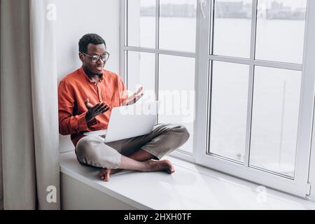 Fröhlicher afroamerikanischer Kerl in stilvoller Kleidung lacht über Videoanruf über einen modernen Laptop, der zu Hause auf einer großen Fensterbank sitzt Stockfoto