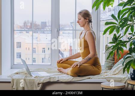 Die blonde junge Frau in gelbem Oberteil und Leggings meditiert in Yoga-Pose und sitzt auf einem mit Decke und Kissen bedeckten Fensterbrett mit einem Laptop Stockfoto