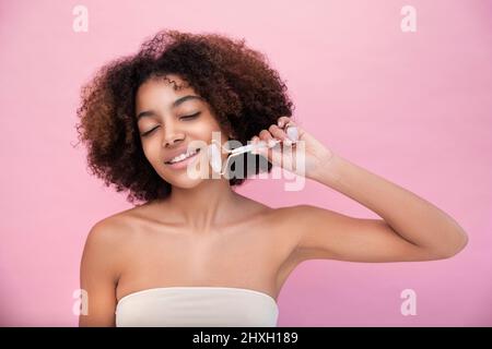 Porträt einer jungen, gepflegten dunkelhäutigen Frau mit üppigen Haaren, die mit Hilfe einer Walze mit geschlossenen Augen auf einem rosa Hintergrund Nahaufnahme, Pflege und Hautpflege eine Gesichtsmassage macht Stockfoto