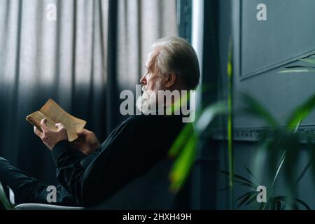 Seitenansicht des konzentrierten älteren grauhaarigen Mannes, der zu Hause ein Papierbuch liest, am Tisch sitzend, entspannt und entspannt sich mit Literatur. Stockfoto