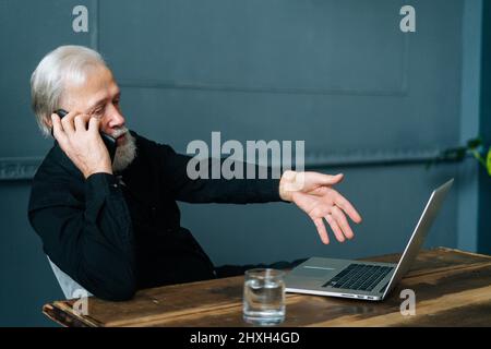 Vorderansicht eines wütenden grauhaarigen Mannes, der mit der technischen Unterstützung sprach und um Hilfe bat, den defekten Laptop-Computer zu reparieren, der am Holztisch sitzt. Stockfoto