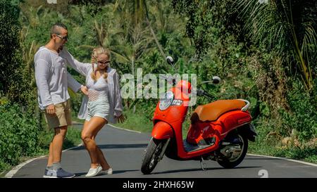 Zwei kaukasische Touristen Mann tanzen in der Nähe von Roller. Liebespaar auf rotem Motorrad in weißer Kleidung, Sonnenbrille auf Waldweg-Tour. Dancing Road. Stockfoto