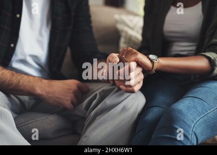 Hand in Hand durch alles. Aufnahme eines unverkennbaren Paares, das die Hände hält, während es zu Hause auf einem Sofa sitzt. Stockfoto