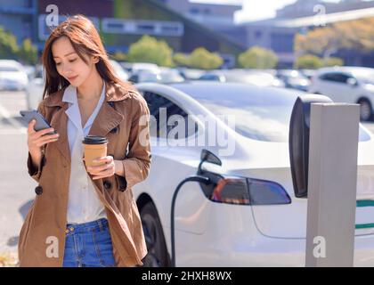 Lächelnde junge Frau, die auf dem Stadtparkplatz in der Nähe eines Elektroautos steht, die Autobatterie von einer kleinen Stadtstation lädt, Kaffee trinkt und smartph benutzt Stockfoto