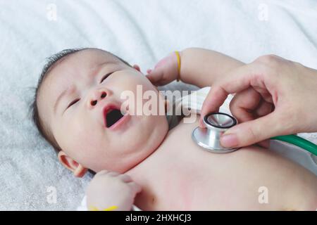 Professioneller Kinderarzt, der das Kleinkind untersucht. Arzt, der ein Stethoskop verwendet, um dem Herzschlag des Kindes zuzuhören. Zwei Monate Baby asiatische Mädchen liegen o Stockfoto