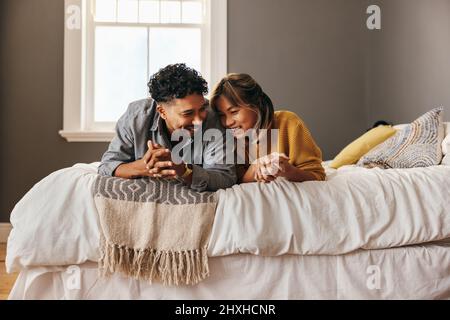 Glückliches interrassisches Paar, das zusammen in ihrem Schlafzimmer lacht. Fröhliches Ehepaar, das sich auf dem Bett entspannen kann. Junges Paar spen Stockfoto