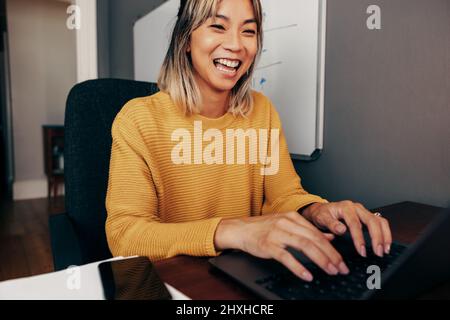 Eine fröhliche Geschäftsfrau tippt eine E-Mail, während sie in ihrem Heimbüro sitzt. Glückliche Unternehmerin lächelt während sie einen Laptop benutzt. Erfolgreiches Geschäftw2 Stockfoto