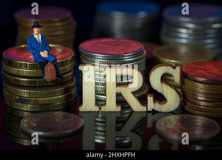 Miniaturmenschen. Mann, der auf Münzstapeln mit Text sitzt. IRS Taxman-Konzept. Stockfoto