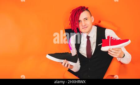Ein lächelnder Schuhverkäufer mit roten Dreadlocks demonstriert Trainer in einem Loch in der Wand. Stilvoller Mann zeigt Turnschuhe, die in den Händen halten. Stockfoto