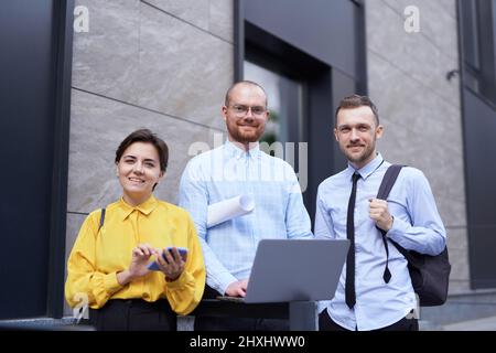 Selbstbewusste Geschäftsleute, die die Kamera betrachten und die Projektpräsentation im Freien besprechen. Team von Mitarbeitern Männer und Frauen in legerer Kleidung im Freien mit Laptop, reden und Brainstorming Stockfoto