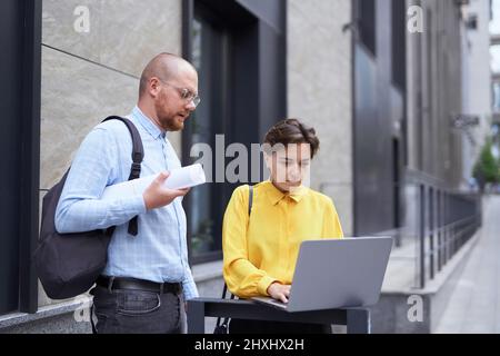 Designer, Architektur- oder Immobilienmanager diskutieren in legerer Kleidung die Projektpräsentation im Freien in der Nähe eines modernen Gebäudes mit Plänen und Laptop. Brainstorming-Konzept. Hochwertige Bilder Stockfoto