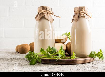 Alterative Kartoffelpflanzenmilch in zwei Glasflaschen auf einem Tablett gegen eine weiße Ziegelwand. Das Konzept einer gesunden vegetarischen Ernährung Stockfoto