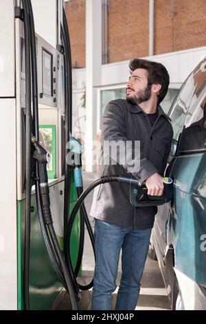 Junger Mann, der sein Fahrzeug betankt, während er sich die Preise an einer Tankstelle ansieht. Stockfoto
