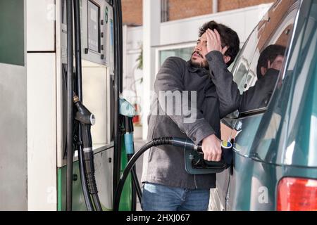 Junger Mann, der sein Fahrzeug betankt, während er sich Sorgen über die hohen Gaspreise macht. Stockfoto