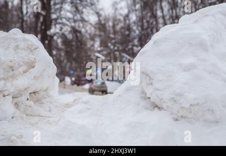 März große Schneeverwehung an der Straße vor dem Hintergrund einer Stadtstraße. Auf der Straße liegt schmutziger Schnee in hohen Haufen. Urbane Winterlandschaft. Wolkiger Wintertag, weiches Licht. Stockfoto