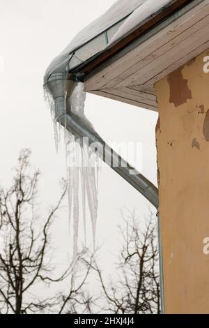 Gefrorene Eiszapfen hängen an einem Abflussrohr am Rand des Dachs. Vor dem Hintergrund des grauen Himmels in der Nähe der Hauswand. Große Kaskaden, sogar schöne Reihen. Wolkiger Wintertag, weiches Licht. Stockfoto