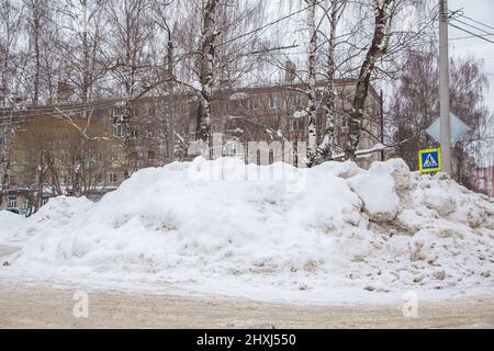 Eine riesige große Schneewehe an der Straße vor dem Hintergrund von Stadthäusern und Bäumen. Auf der Straße liegt schmutziger Schnee in hohen Haufen. Urbane Winterlandschaft. Wolkiger Wintertag, weiches Licht. Stockfoto