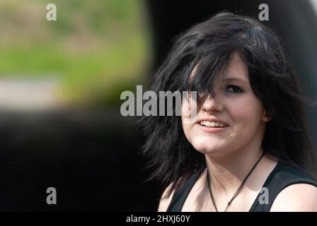 Punk-Emo-Mädchen, junger Erwachsener mit schwarzen Haaren und Eyeliner, lächelnd, Blick auf die Kamera, im Freien, horizontal, Nahaufnahme Stockfoto