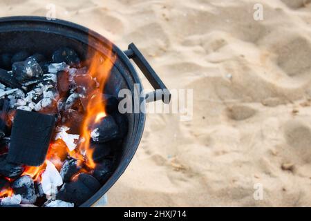 Nahaufnahme von Holzkohle mit einer gelben Flamme in einem runden metallischen Grill an einem Sandstrand Stockfoto