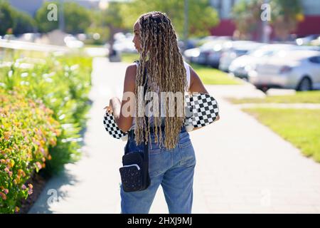 Schwarze Frau mit farbenfroher geflochtener Frisur, die ein Skateboard trägt. Stockfoto