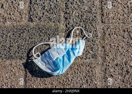 Gebrauchte blaue Gesichtsmaske, die auf dem Boden auf einer ländlichen Kopfsteinpflasterstraße liegt, Umweltverschmutzung in Zeiten der Ausbreitung von Covid-19 oder des Coronavirus-Ausbruchs Stockfoto