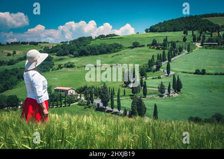 Ziemlich fröhliche Frau in einem roten Rock weißen Hemd und einem Strohhut genießen die Aussicht in der Toskana. Berühmter ländlicher touristischer Ort mit Getreidefeldern und Curv Stockfoto