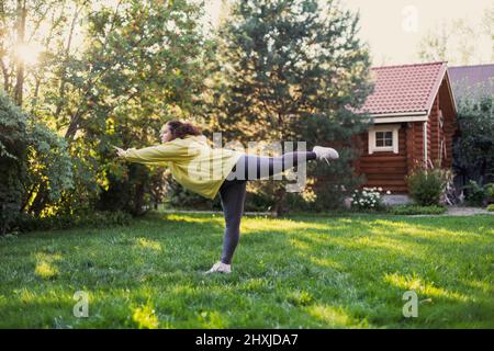 Kaukasische fette Frau, die auf einer Hand beim Yoga draußen steht und Sportkleidung auf grünem Gras auf dem Hinterhof mit Landhaus und hohen Bäumen trägt Stockfoto