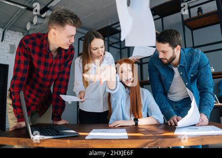 Wütend erschöpfte junge Geschäftsfrau unter Stress warf Dokumente am Schreibtisch sitzend auf die Kamera, multirassische Kollegen in ihrer Nähe stehen. Stockfoto