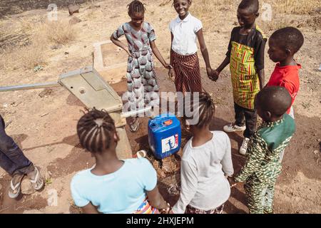 Gruppe afrikanischer Jungen und Mädchen, die auf die Befüllung eines Wassertanks am Dorfbrunnen warten; fehlende Wasserinfrastrukturen in der subsaharischen Region Stockfoto