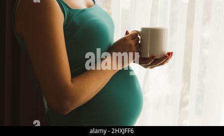 Mug Mockup. Schwangere mit weißem Becher. Schwangere trinkt Kaffee am Fenster. 6 Monate schwanger Frau trinken heißen Tee stehend Stockfoto