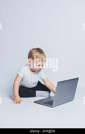 Ein kleiner Junge, 3-4 Jahre alt, sitzt mit einem Laptop auf weißem Hintergrund. Ein Kind in einem weißen T-Shirt und einer schwarzen Hose schaut auf einen Laptop. Moderne Kinder Stockfoto