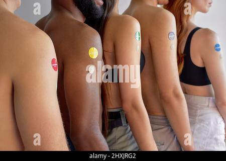 Junge Menschen stehen in der Schlange, nach der Impfung in der Schlange. Drinnen. Seitenansicht, Nahaufnahme der Arme. Kurzgeschnittene, nicht erkennbare Männer und Frauen mit unterschiedlicher Haut Stockfoto