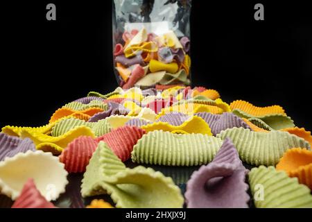 Kreative bunte Pasta-Farfalle auf schwarzem Hintergrund in Perspektive mit Verpackung im Hintergrund. Extremes Makro Stockfoto