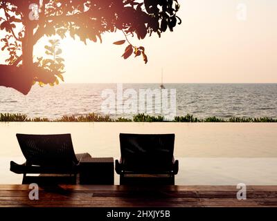 Zwei einsame, leere moderne Rattanliegen, Sessel mit Beistelltisch auf dem Holzdeckenboden und der Pool unter dem Baum auf dem friedlichen Meer Stockfoto
