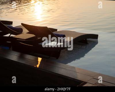 Zwei leere, moderne Rattanliegen, Liegestühle für die Gäste auf weißem Marmorboden am Pool im Hotelresort auf friedlichem Hintergrund am Himmel bei Sonnenuntergang mit Kopien Stockfoto