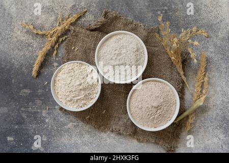 Lebensmittel und Backzutat. Weizenmehl grob aus Vollkorngetreide, Weizenkleie und Weizenmehl auf grauem Steingrund. Draufsicht flach liegend. Stockfoto