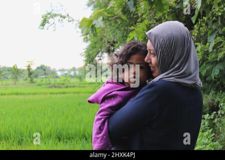 Java, Indonesien - 12 24 2020: Verschwommene Hintergrundansicht von Reisfeldern und einer Frau in einem Hijab, die ein Mädchen hält, sieht glücklich aus Stockfoto