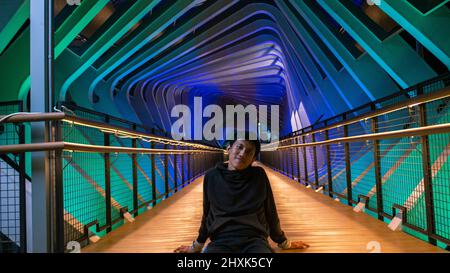 Männer, die lässig im Korridor der Fußgängerbrücke sitzen, die mit bunten Lichtern geschmückt ist und nachts einsam aussieht Stockfoto